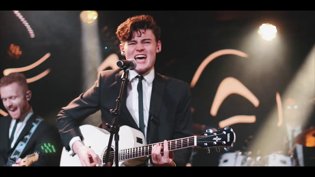 Young male musician passionately singing and playing a white acoustic guitar on stage with a band, wearing a suit and tie.