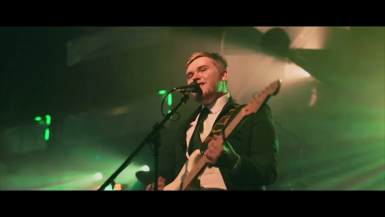 Musician in suit playing electric guitar and singing into microphone on stage with green lighting.