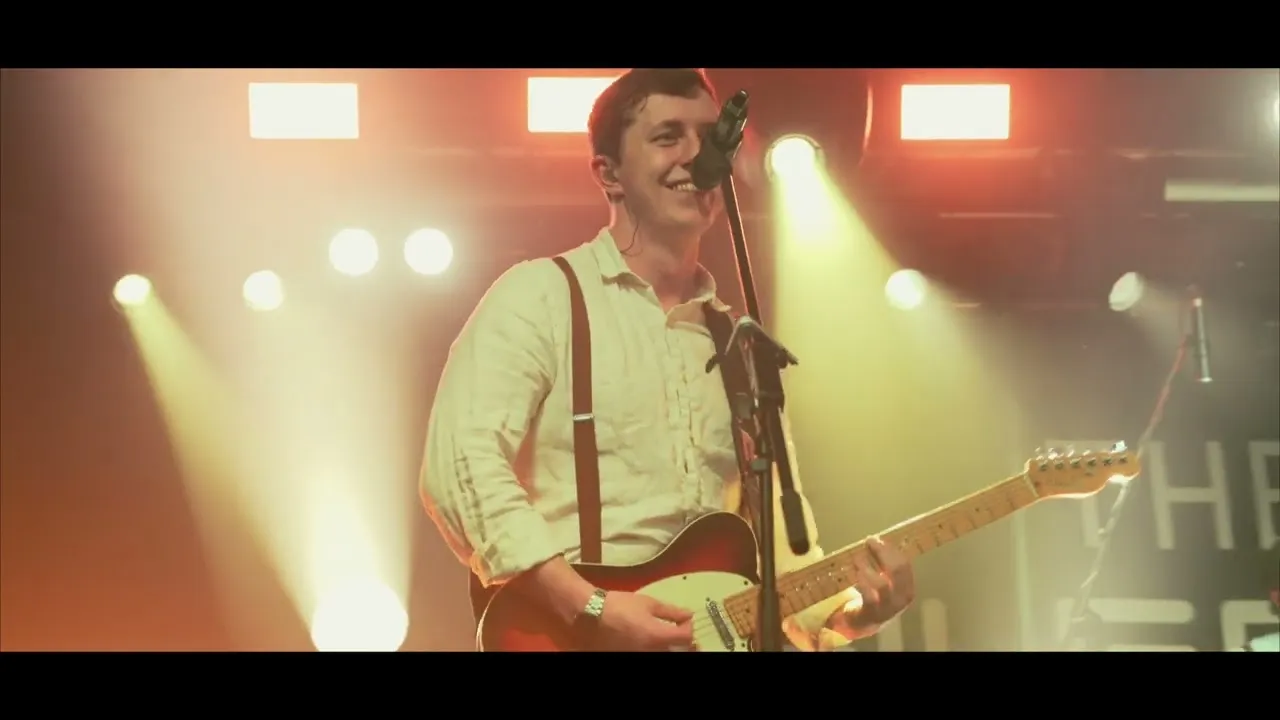 Smiling musician playing electric guitar and singing into a microphone on stage with warm yellow and orange stage lights.