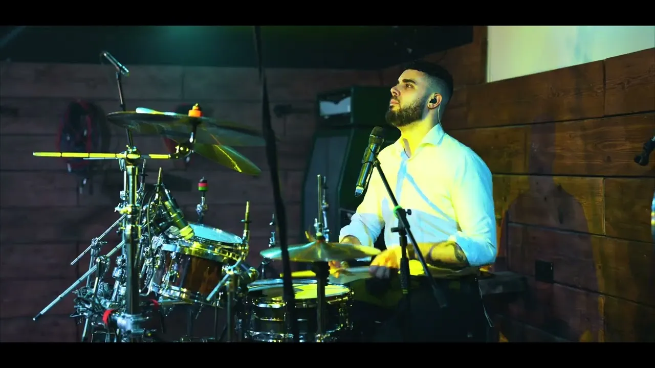 Bearded man in a white shirt playing drums in a wooden room with microphone setup.