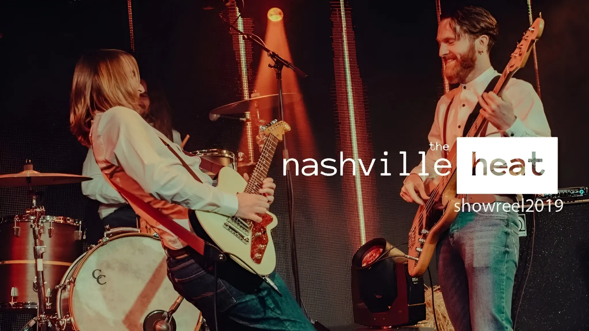 Two musicians playing guitar and bass on stage with a drummer in the background, under warm stage lighting.