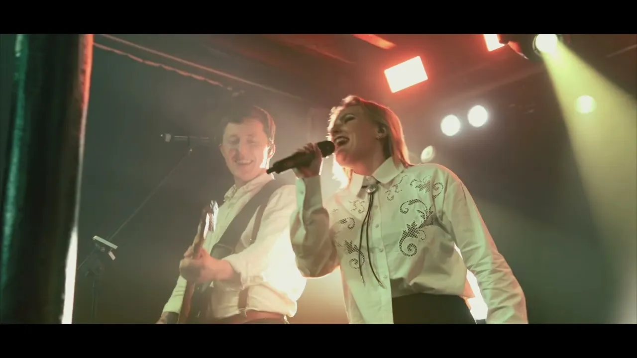A male guitarist and a female singer performing on stage with bright stage lights behind them.