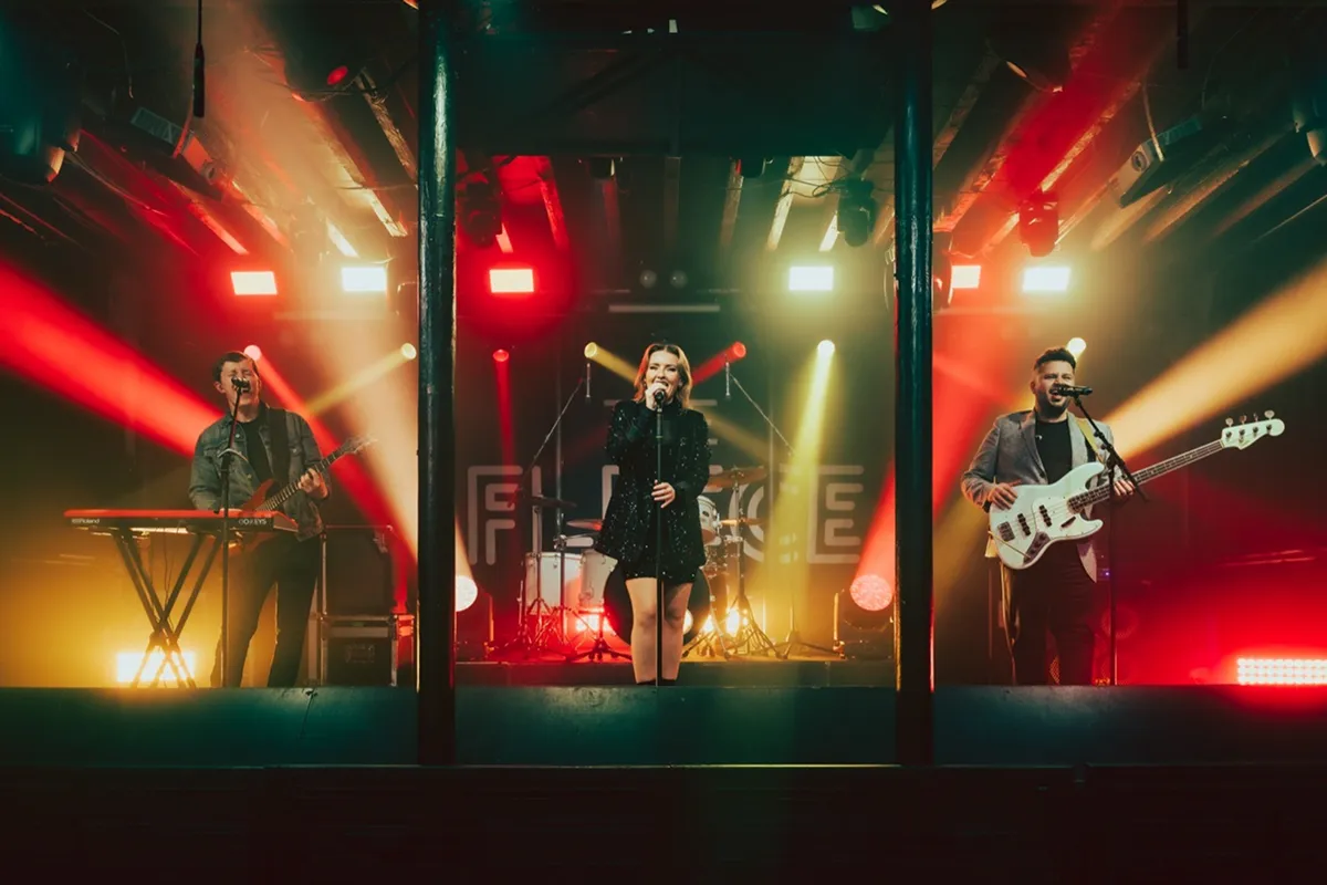 Three musicians performing on stage with colorful red and yellow lights and a drum set in the background.