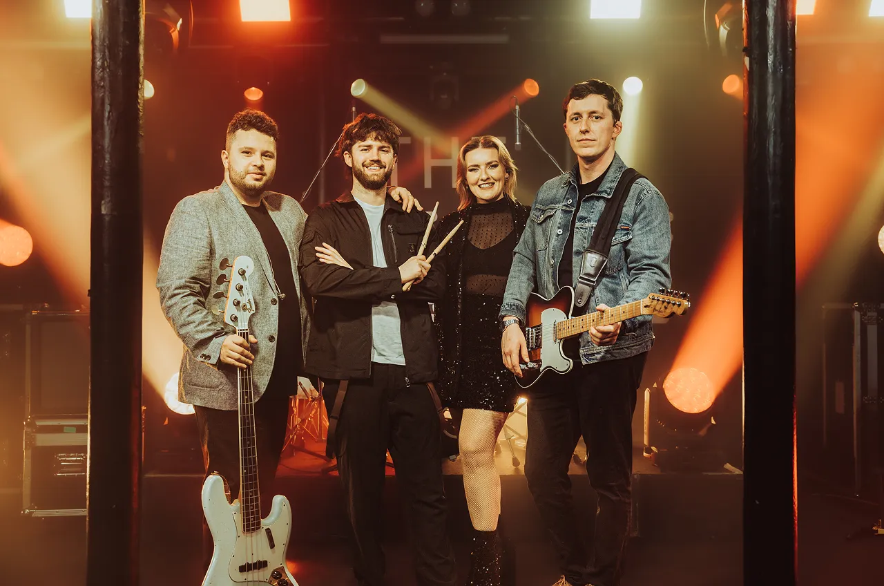 Four musicians posing on stage with instruments including a bass guitar, electric guitar, and drumsticks under warm stage lighting.