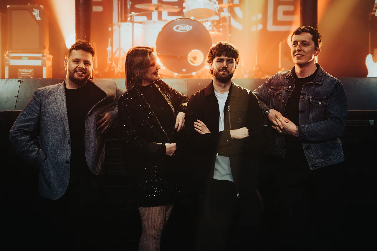 Four band members standing in front of a drum set on a stage with warm orange lighting.