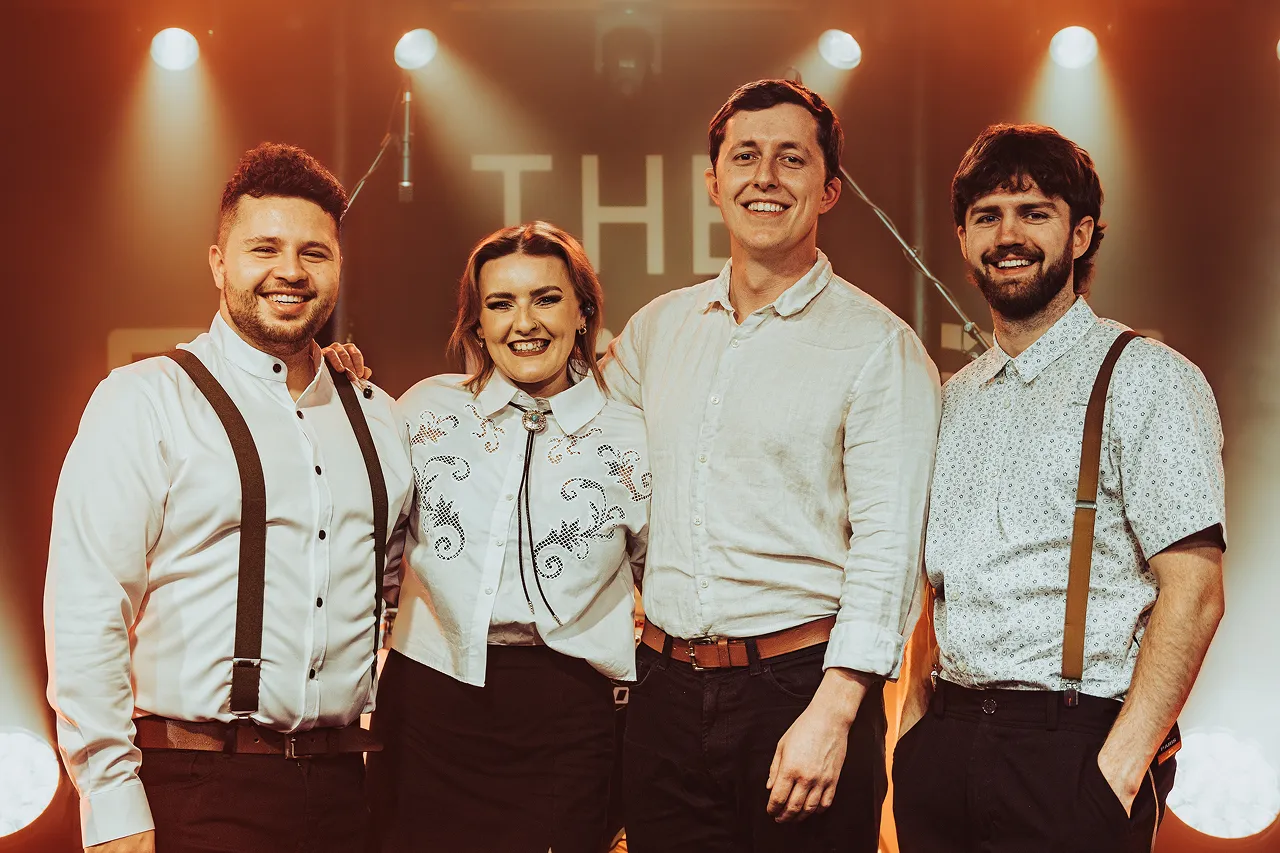 Four smiling band members standing together on stage under warm spotlights.