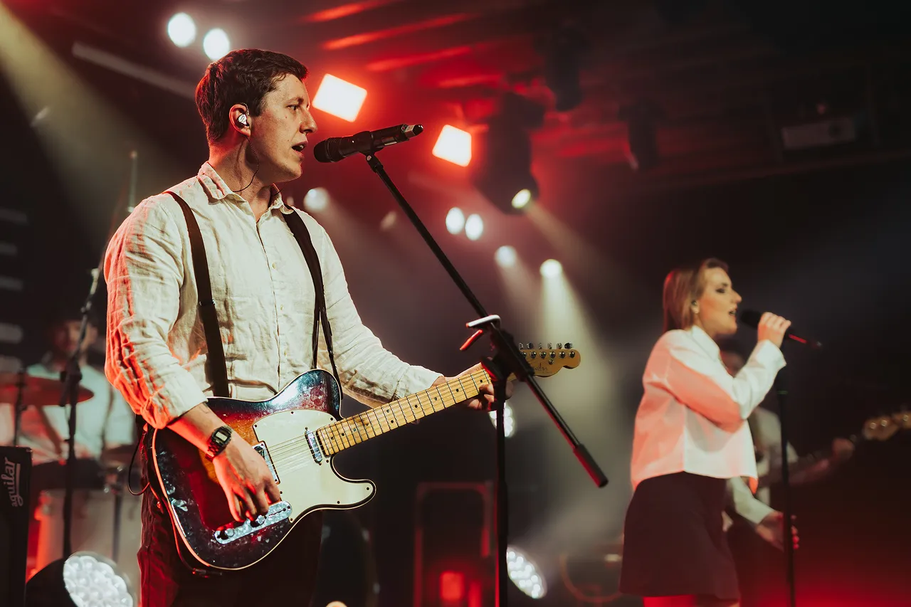 Male guitarist singing into a microphone and playing electric guitar on stage with female singer in background under red and white stage lights.