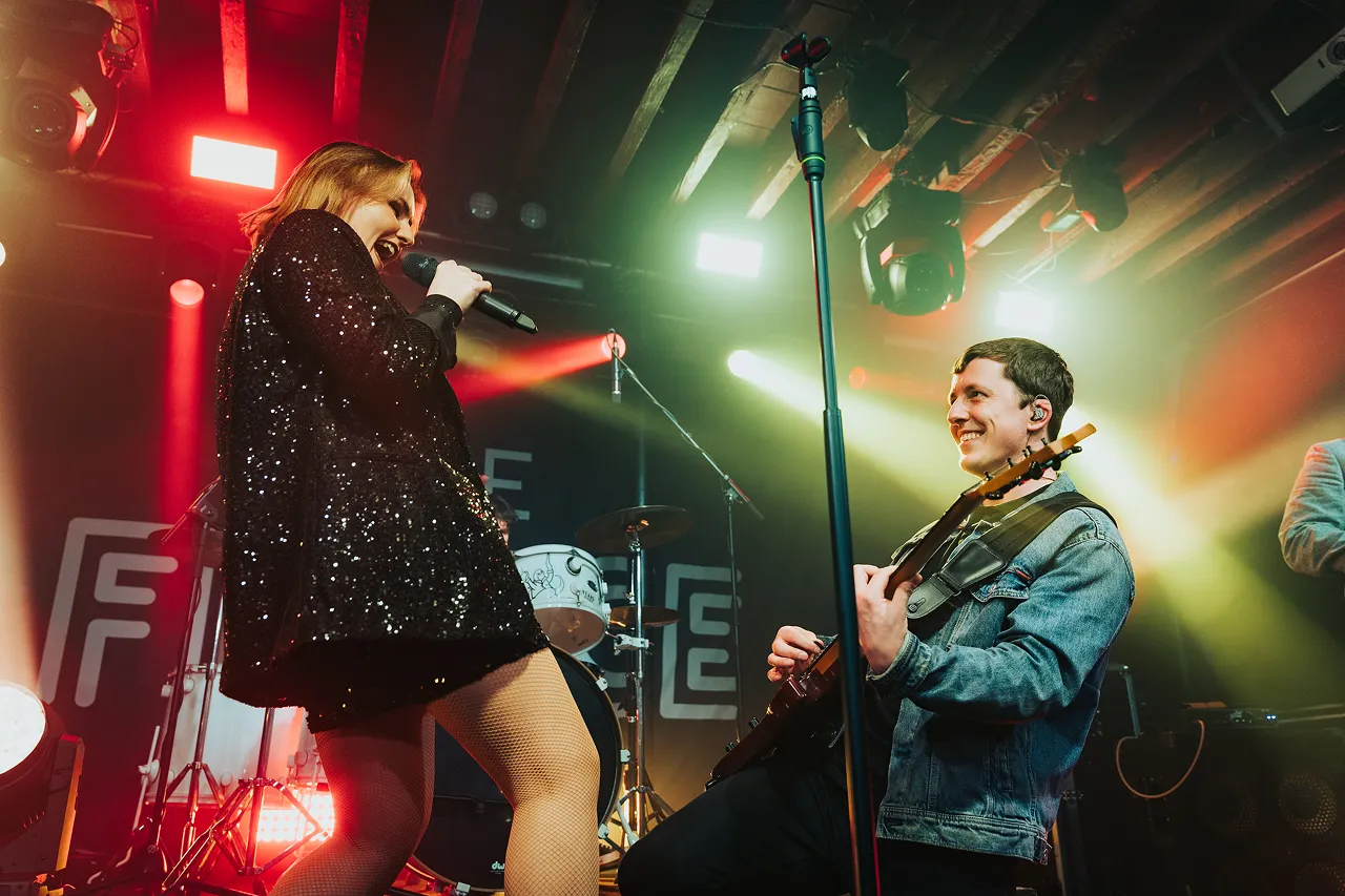 Female singer in a sparkling black outfit interacting energetically with a smiling male guitarist on stage under colorful stage lights.