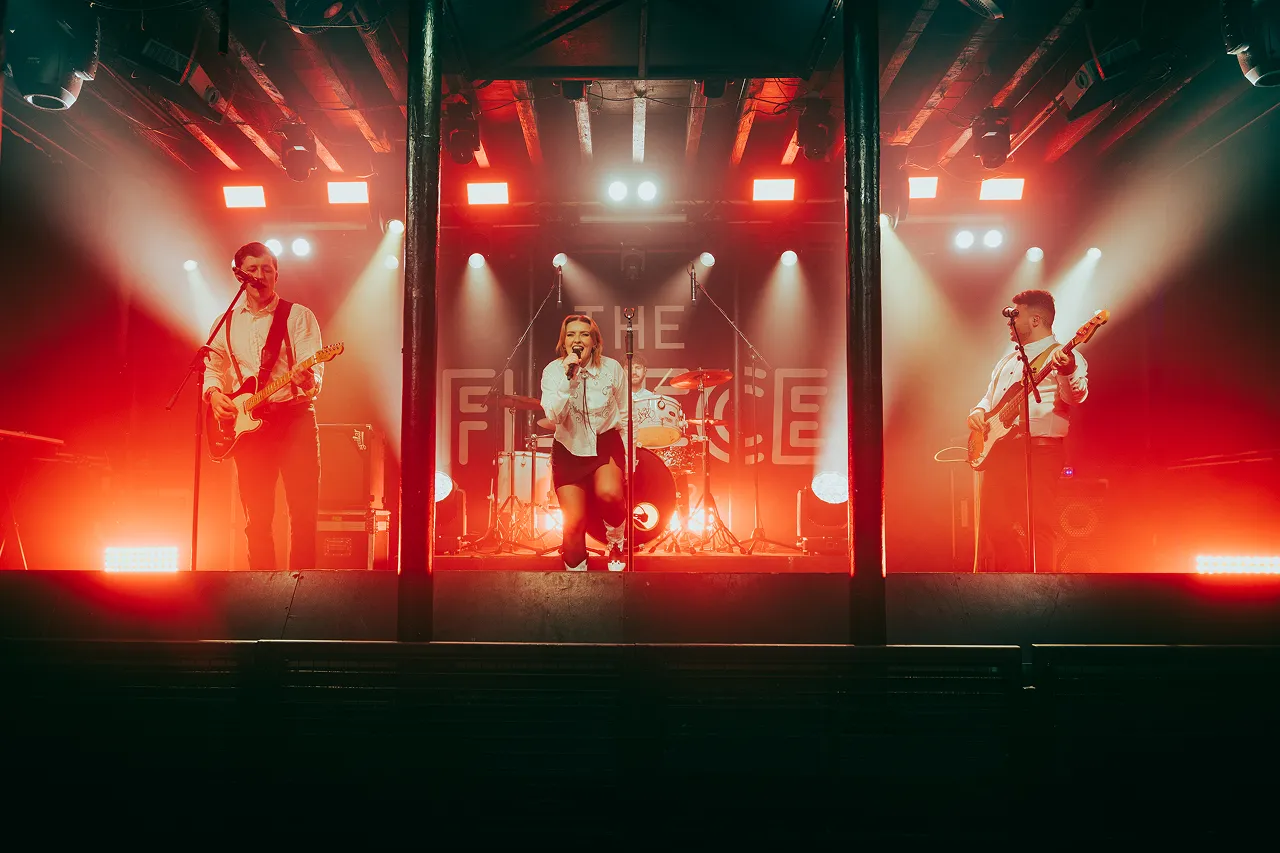 A four-member band performing on stage with red and white lighting, featuring a female vocalist kneeling at the center, a guitarist on the left, a drummer in the back, and a bassist on the right.