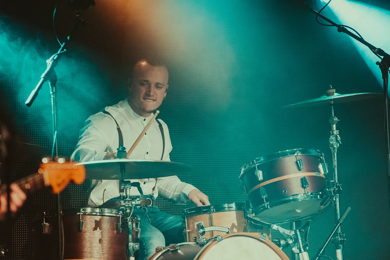 Male drummer playing drum set on stage under blue and orange lighting.