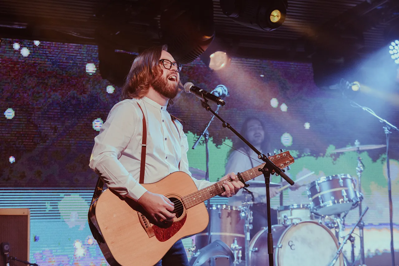 Bearded musician with glasses playing acoustic guitar and singing into microphone on stage with drummer in background.
