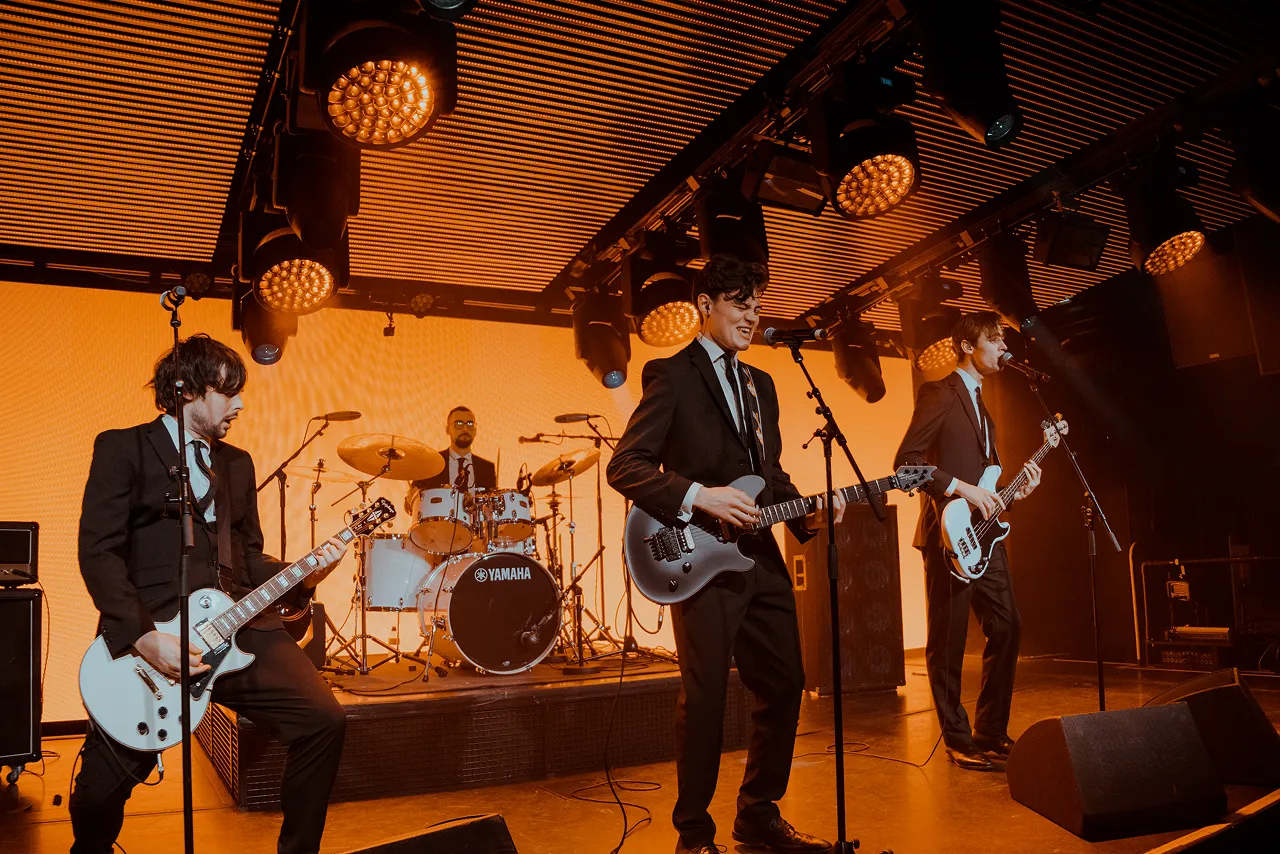 Four-member band performing on stage with guitars and drums under warm orange stage lighting.