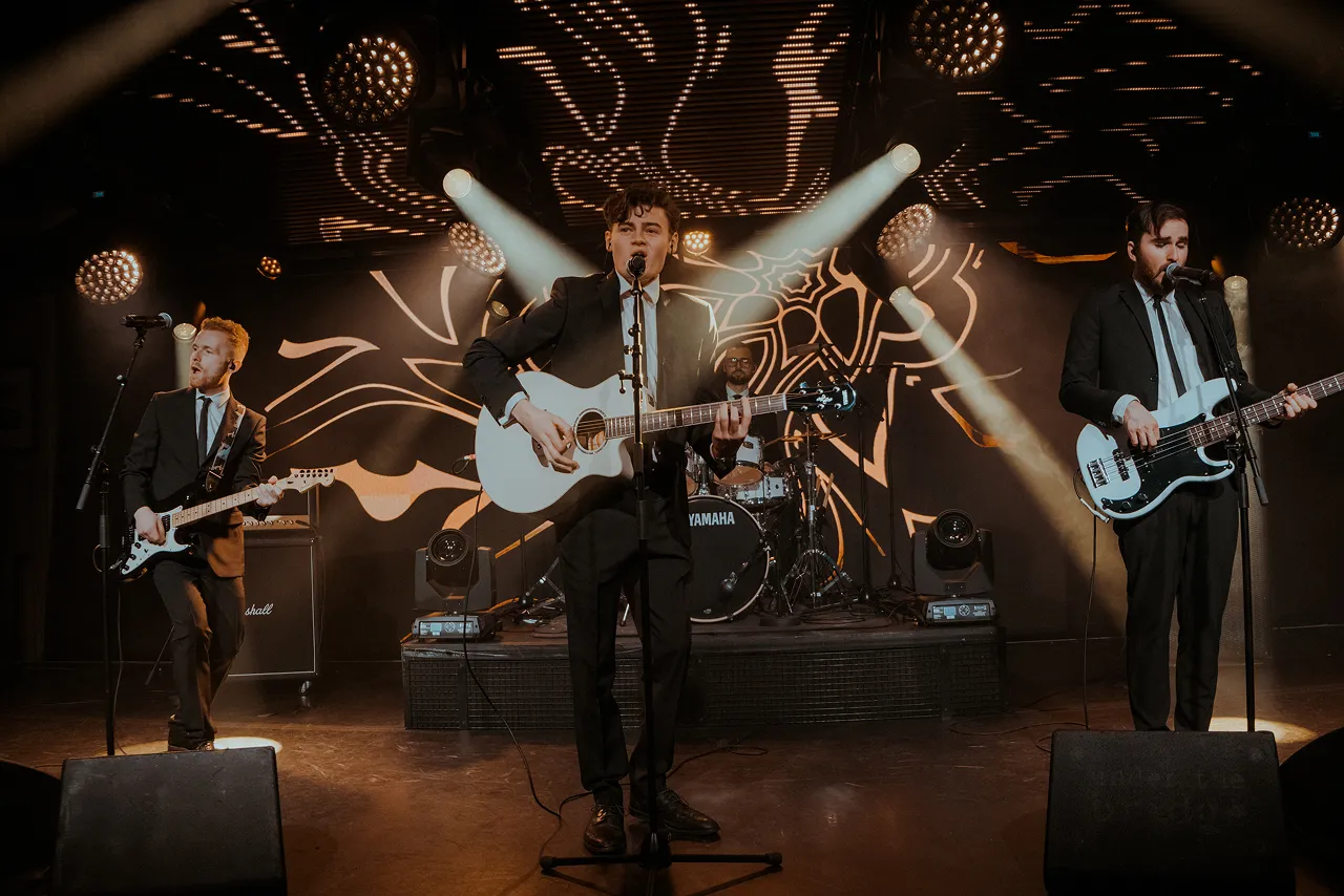 Four-piece band wearing suits performing on stage with guitars and drums under spotlights.