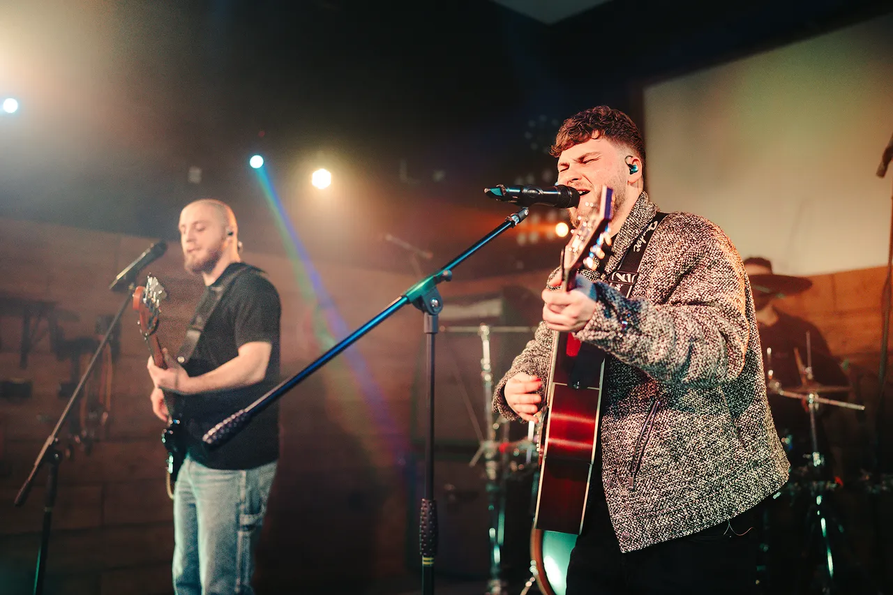Two musicians performing on stage with one singing and playing an acoustic guitar and the other playing an electric guitar.
