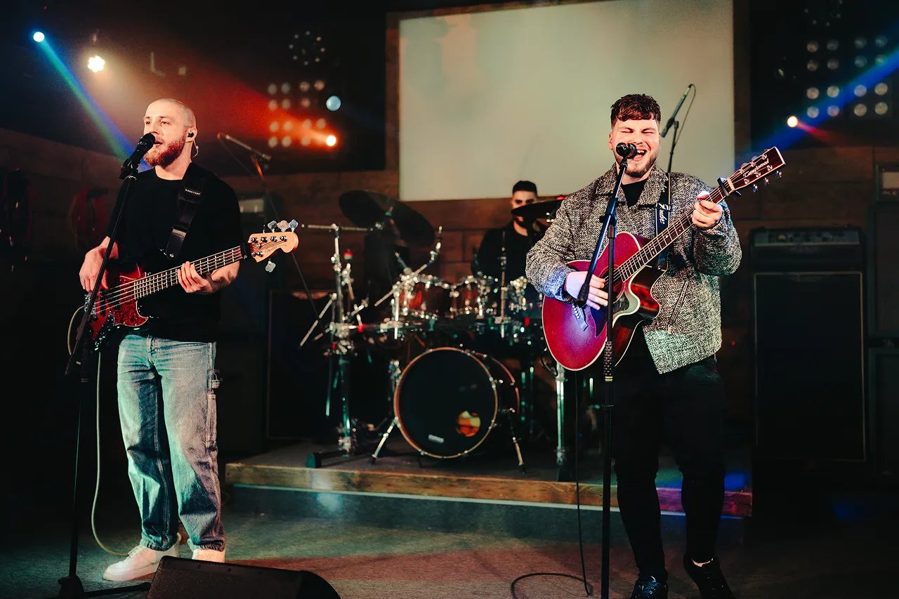 Three musicians performing on stage with guitar, bass, and drums under colorful stage lights.